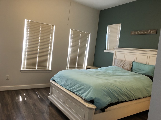 A bedroom with a bed, two windows with blinds, and a wooden floor.