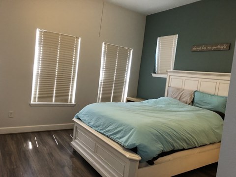 A bedroom with a bed, two windows with blinds, and a wooden floor.