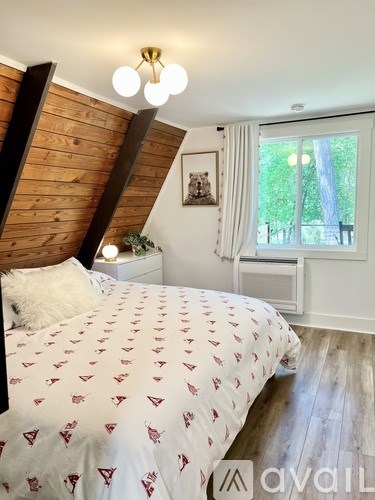 A bedroom with a bed covered in a white comforter with red triangles, a wooden headboard, and a window with white curtains.