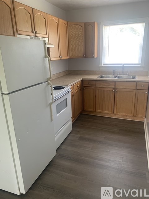 A white refrigerator stands in a kitchen with wooden cabinets and a window.