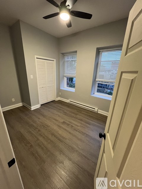 A room with a ceiling fan and light, wooden flooring, and two windows with blinds.
