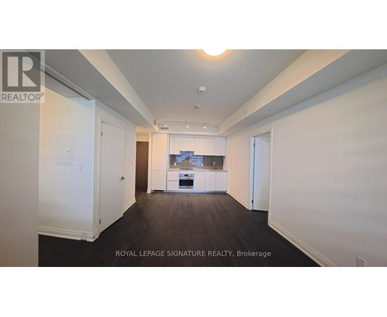 A white room with a kitchen in the background and a brokerage sign on the wall.