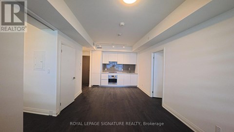 A white room with a kitchen in the background and a brokerage sign on the wall.