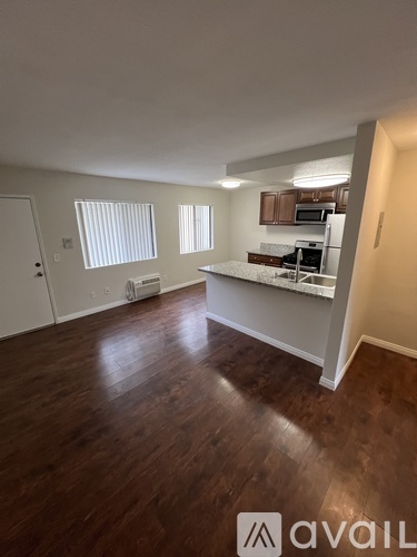 A room with wooden floors and a kitchen area with white cabinets and appliances.