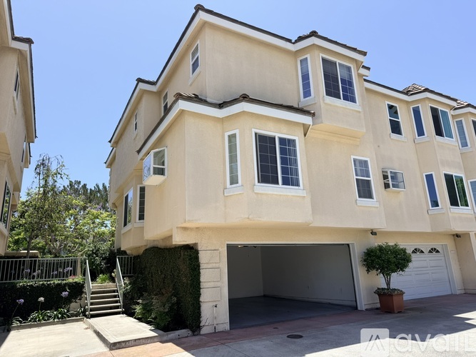 A two-story beige house with a garage and a small tree in front.
