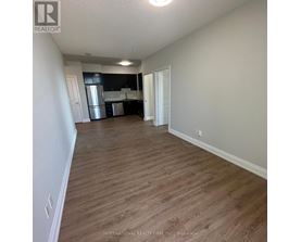 A room with wooden flooring and white walls, with a kitchen area visible in the background.