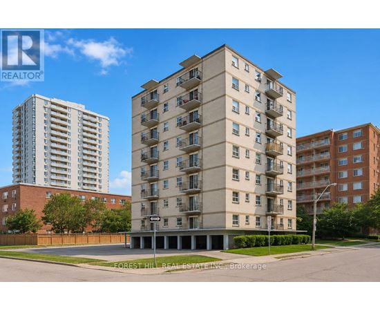 A tall beige apartment building stands in front of a smaller building and a tree.