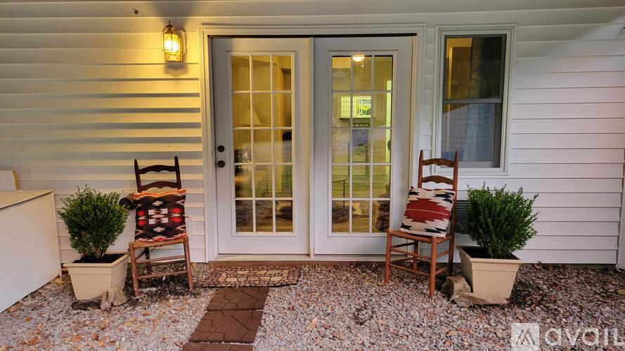 A white house with a glass door and a small porch with two chairs and two potted plants.