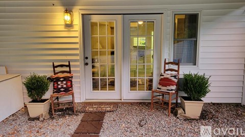 A white house with a glass door and a small porch with two chairs and two potted plants.