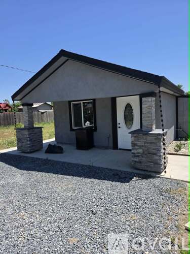 A small house with a grey roof and a black door is surrounded by a gravel driveway.