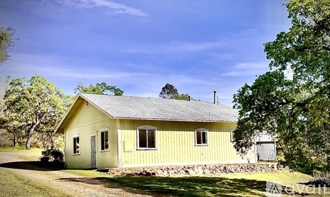 A yellow house with a grey roof and a chimney is surrounded by greenery.
