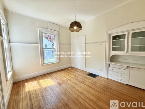 A room with wooden flooring and white walls, with a window showing a red building outside.