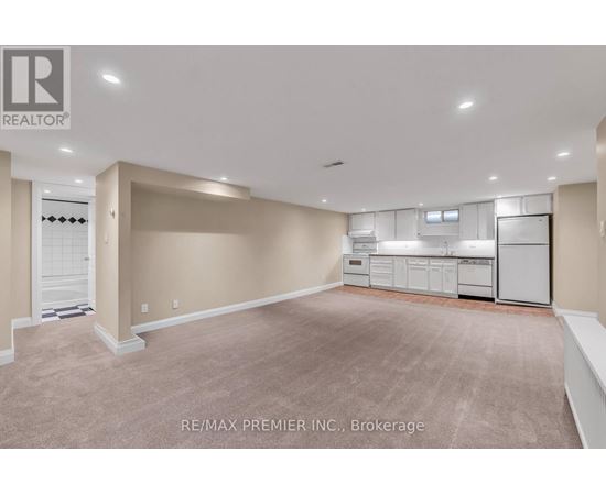 A spacious room with beige carpeting and white walls, featuring a kitchenette with white appliances.