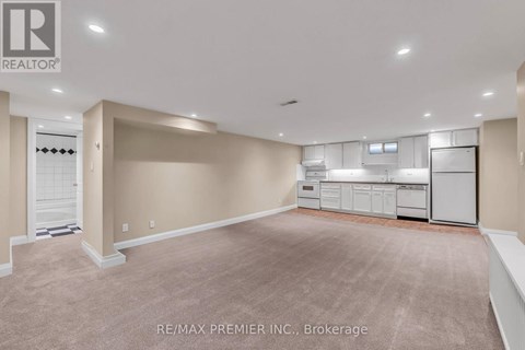 A spacious room with beige carpeting and white walls, featuring a kitchenette with white appliances.