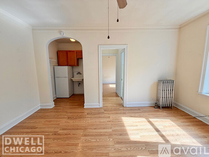 A room with wooden flooring and white walls, with a kitchen visible through an archway.