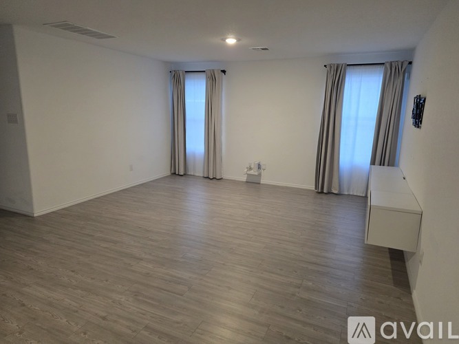 A spacious room with wooden flooring and two windows with curtains.