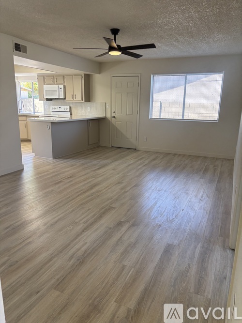 A room with a ceiling fan and light, wooden floors, and a kitchen area with white cabinets and appliances.