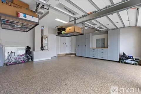 A spacious room with a grey floor and white walls, featuring a kitchen area with white cabinets and a countertop, a large window with a view of the sky, and a few pieces of furniture and decorations scattered throughout the space