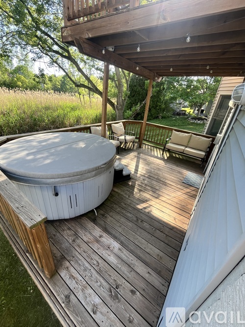 A hot tub sits on a wooden deck with a white couch and a wooden pergola above it.