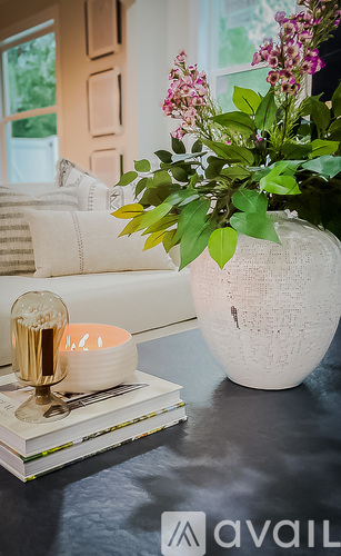 A white vase with green leaves and pink flowers sits on a table next to a stack of magazines and a candle holder.