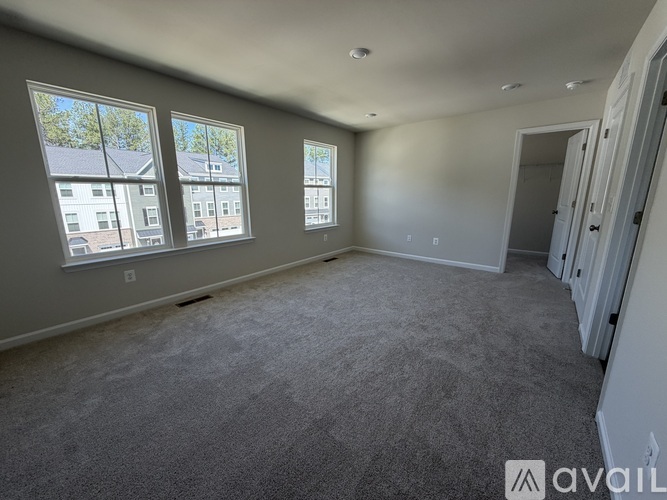 A spacious room with grey carpeting and three windows showing a view of buildings outside.