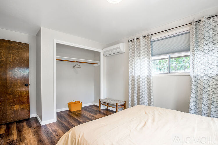 A bedroom with a bed, a window with curtains, a wooden door, and a small table with a chair.