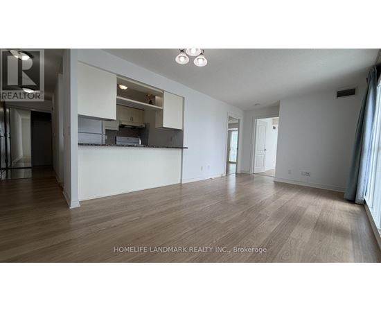 A spacious room with wooden flooring and white walls, with a kitchen area visible through a glass door.