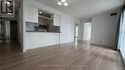 A spacious room with wooden flooring and white walls, with a kitchen area visible through a glass door.