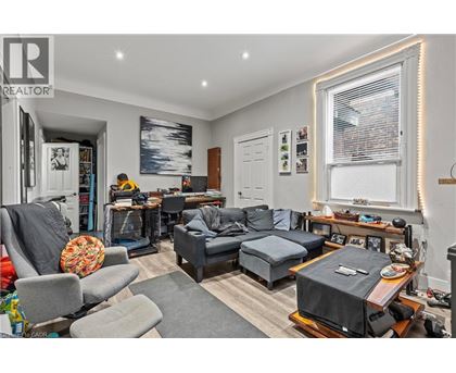 A living room with a grey couch, a grey armchair, a grey ottoman, a wooden coffee table, a TV on a stand, a bookshelf, a window with blinds, a painting on the wall, and a