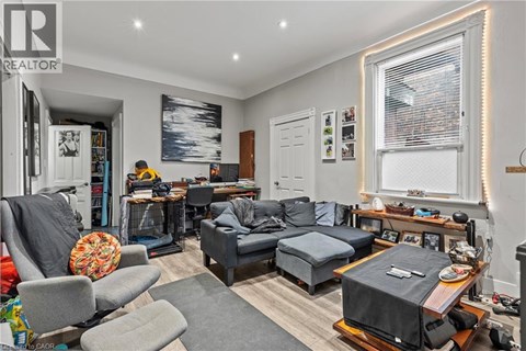 A living room with a grey couch, a grey armchair, a grey ottoman, a wooden coffee table, a TV on a stand, a bookshelf, a window with blinds, a painting on the wall, and a