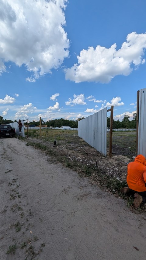 A man in an orange jacket is crouched down beside a metal fence.