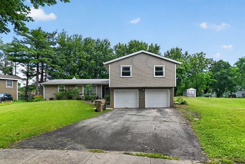 A house with a grey garage door is surrounded by green grass and trees.