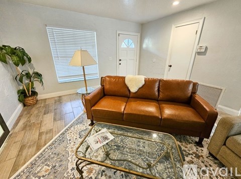 A brown leather couch with a white pillow is in the center of a living room with a glass coffee table in front of it.
