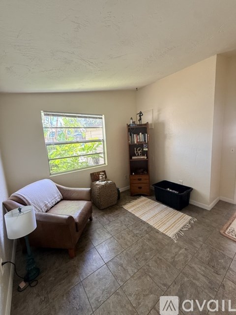 A living room with a brown leather couch, a white lamp, a window with a view of greenery outside, a bookshelf with various items, and a rug on the floor.