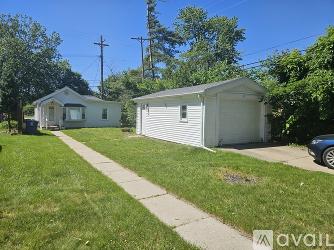 A white house with a garage is situated in a grassy area with a sidewalk leading to it.