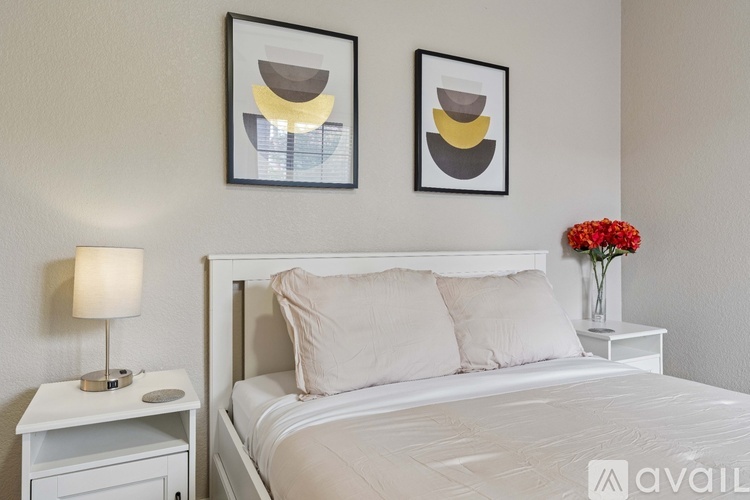 A bedroom with a white bed, a white nightstand with a lamp, and two framed artworks on the wall.