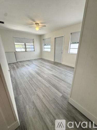 A spacious room with a ceiling fan and light, hardwood flooring, and a large window.
