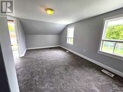 A room with grey carpeting and a window showing greenery outside.