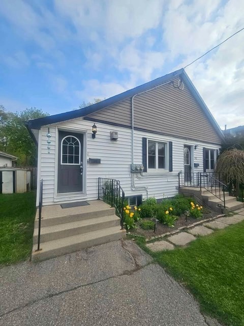 A house with a grey siding and a black door is surrounded by a small garden with yellow flowers.