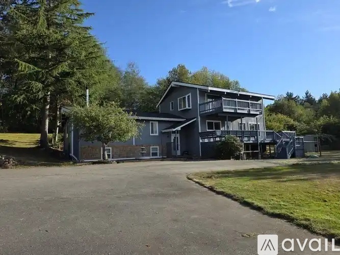 A large house with a balcony is surrounded by trees and has a driveway in front.