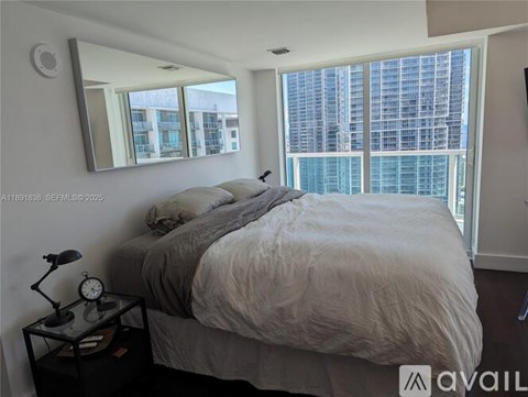A bedroom with a large bed, a nightstand with a clock, and a large window with a view of a city.