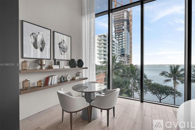 A modern dining room with a glass table and chairs, a shelf with decorative items, and a view of a cityscape through the window.