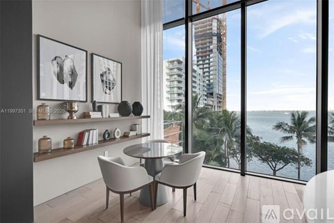 A modern dining room with a glass table and chairs, a shelf with decorative items, and a view of a cityscape through the window.