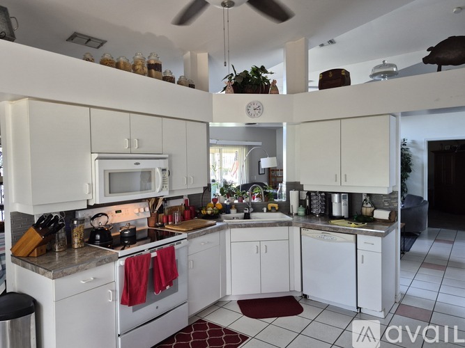 A kitchen with white cabinets and appliances, a red rug on the floor, and a ceiling fan.