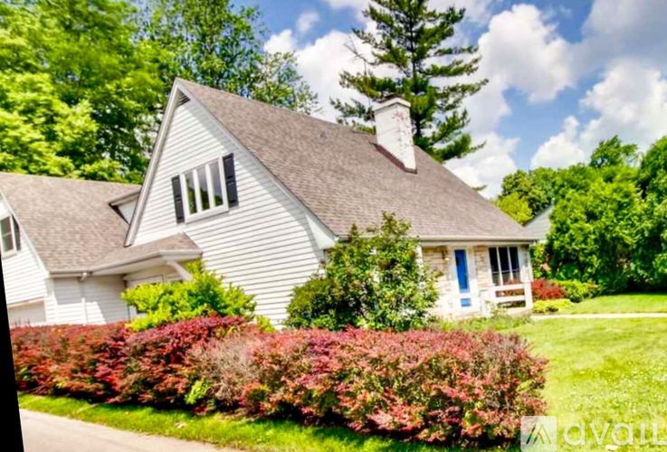 A white house with a brown roof and a chimney is surrounded by greenery.