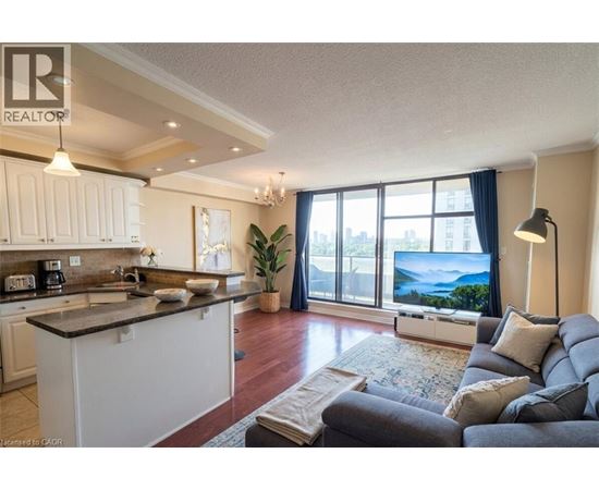 A well-lit, spacious living room with a grey sofa and a kitchen area with white cabinets.
