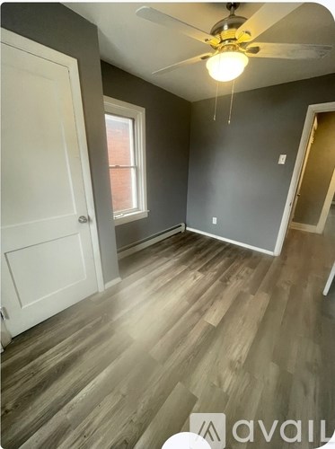A room with a ceiling fan and light, wooden flooring, and a window with a view of a red building outside.
