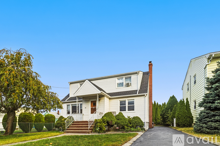 A house with a white exterior and a red brick chimney is surrounded by greenery and a clear blue sky.