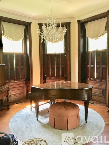 A grand piano is in a room with a chandelier and a round ottoman.