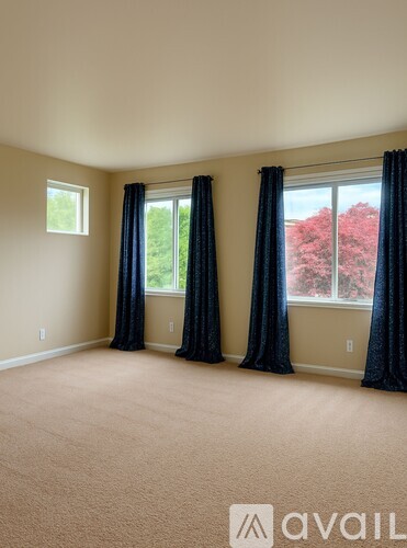A room with a carpeted floor, two windows with black curtains, and a view of red trees outside.